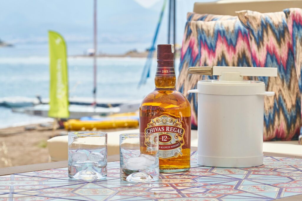 Chivas Regal whiskey bottle with ice glasses by the beach, perfect for a relaxing summer evening.